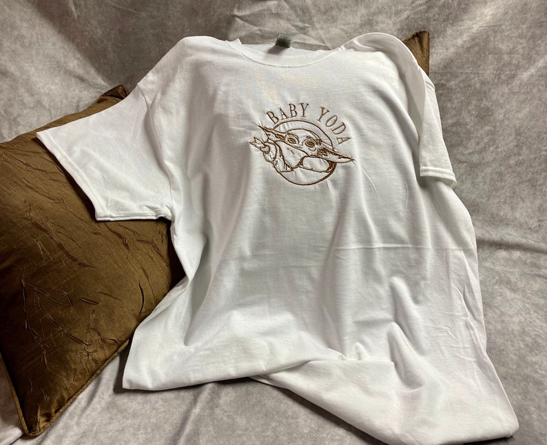 May include: White t-shirt with a brown embroidered image of Baby Yoda and the text "Baby Yoda".
