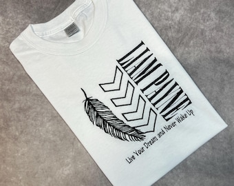 May include: White t-shirt with a black graphic design that says "I am Pain" with a feather and arrow design. The text below the graphic says "Live Your Dream and Never Wake Up".