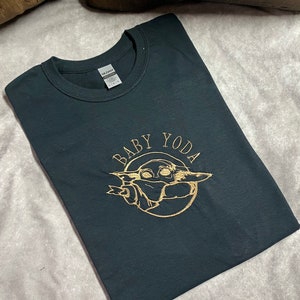 May include: Black t-shirt with a gold embroidered image of Baby Yoda, surrounded by a circle with the text "BABY YODA".