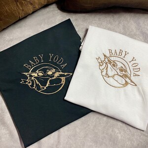 May include: Two t-shirts with a gold embroidered image of Baby Yoda. The text "Baby Yoda" is also embroidered in gold on both shirts. One shirt is black and the other is white.