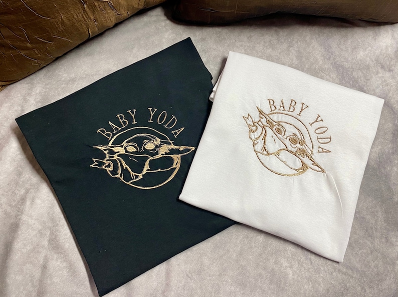 May include: Two t-shirts with a gold embroidered image of Baby Yoda. The text "Baby Yoda" is also embroidered in gold on both shirts. One shirt is black and the other is white.