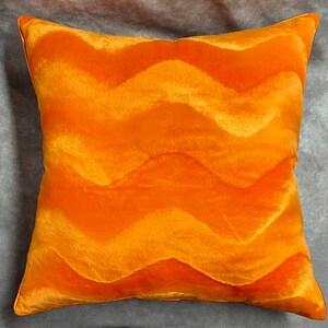 K&ouml;nnte beinhalten: Orangefarbener Dekokissen mit wellenf&ouml;rmigem Muster. Das Kissen ist aus weichem, pl&uuml;schem Stoff.