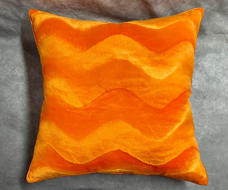 K&ouml;nnte beinhalten: Orangefarbener Dekokissen mit wellenf&ouml;rmigem Muster. Das Kissen ist aus weichem, pl&uuml;schem Stoff.