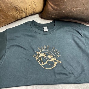 May include: A dark gray t-shirt with a gold graphic of Baby Yoda. The graphic is a circle with the words "Baby Yoda" around the outside and a picture of Baby Yoda inside.