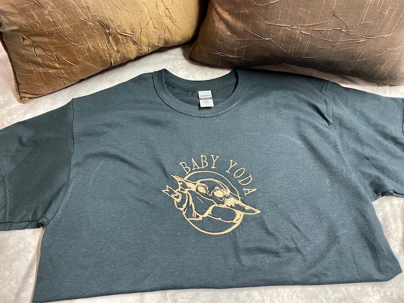 May include: A dark gray t-shirt with a gold graphic of Baby Yoda. The graphic is a circle with the words "Baby Yoda" around the outside and a picture of Baby Yoda inside.