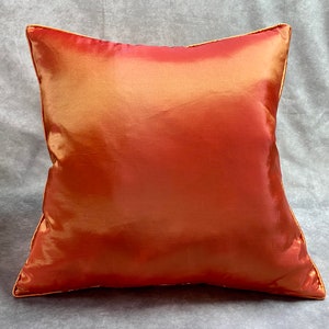K&ouml;nnte beinhalten: Ein quadratisches Kissen mit einem weichen, seidenartigen, orangefarbenen und goldenen Stoff. Das Kissen hat einen dezenten Glanz und eine glatte Textur.