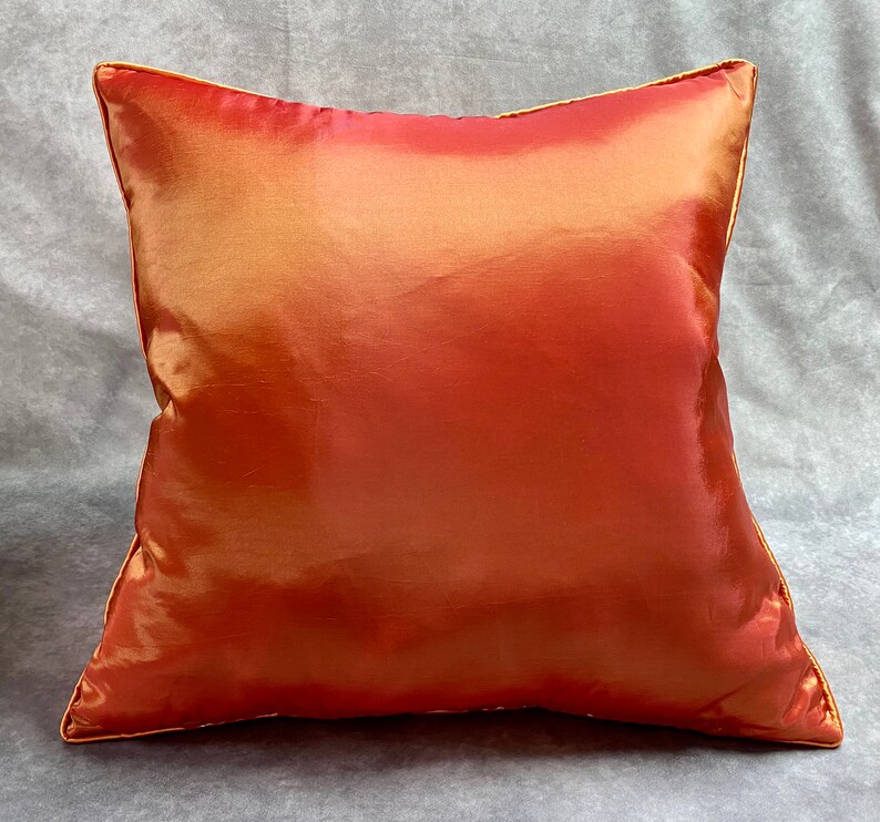 K&ouml;nnte beinhalten: Ein quadratisches Kissen mit einem weichen, seidenartigen, orangefarbenen und goldenen Stoff. Das Kissen hat einen dezenten Glanz und eine glatte Textur.