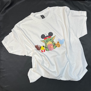 May include: White t-shirt with a green character wearing Mickey Mouse ears and holding a bucket of popcorn. The character is surrounded by other Disney-themed icons, including a Mickey Mouse ice cream cone, a Mickey Mouse balloon, and a brown character holding a Mickey Mouse ice cream bar.