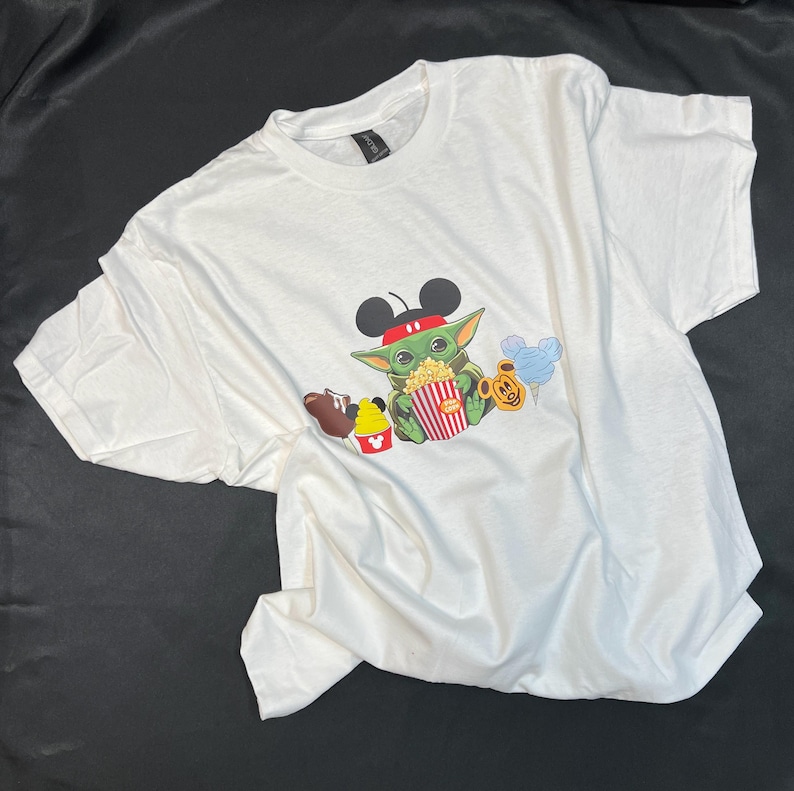 May include: White t-shirt with a green character wearing Mickey Mouse ears and holding a bucket of popcorn. The character is surrounded by other Disney-themed icons, including a Mickey Mouse ice cream cone, a Mickey Mouse balloon, and a brown character holding a Mickey Mouse ice cream bar.