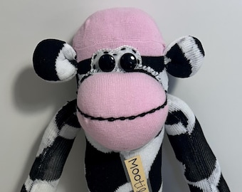 May include: A handmade sock monkey doll with a pink face, hat, and feet. The monkey's body is white with black spots. The doll has black button eyes and a stitched mouth. A small tag reads "MooHilda".