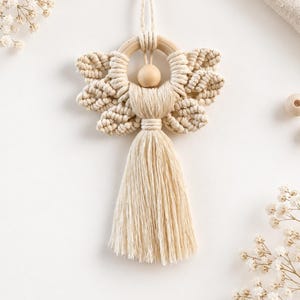May include: A macrame angel ornament crafted from off-white cotton cord, featuring a circular wooden ring halo, a smooth wooden bead head, and intricate knotted wings. The figure includes a long, flowing tassel skirt and a looped cord for hanging.