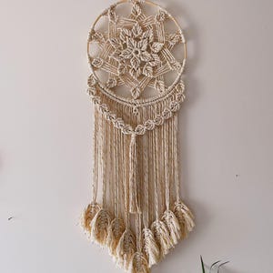 Op de afbeelding: Een crèmekleurige macramé wandhanger. Het cirkelvormige ontwerp heeft een centraal sterrenpatroon en een waterval van franjes. Het stuk is gemaakt van geweven natuurlijke vezels en heeft een bohemien uitstraling.