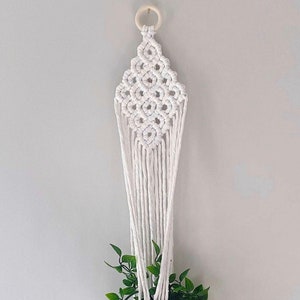 May include: White macrame plant hanger with a wooden ring at the top. The hanger is in the shape of a diamond and has a plant pot hanging from it. The text "PDF PATTERN" and "STEP-BY-STEP VIDEO TUTORIAL INCLUDED" is at the bottom of the image.