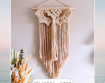 Puede incluir: Un tapiz de macramé beige con un patrón geométrico y flecos. El tapiz está sujeto a una varilla de madera. "PDF EBOOK + VIDEO" está escrito en negro en la parte inferior de la imagen.