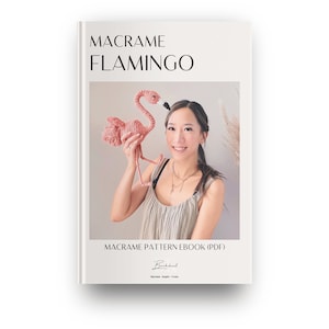 Può includere: Una copertina di libro bianca con il titolo "Macrame Flamingo" e il testo "Macrame Pattern Ebook (PDF)". La copertina presenta una foto di una donna che tiene un fenicottero rosa in macramè.