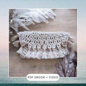 May include: A white macrame clutch bag with a beaded design and fringe trim. The bag is on a wooden surface with dried foliage.
