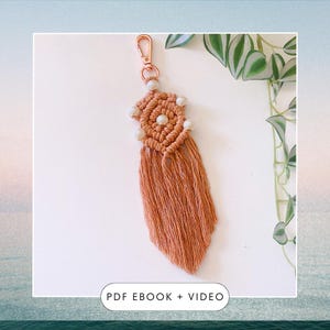 Peut inclure: Un porte-clés en macramé de couleur pêche avec un motif en forme d'étoile et des perles blanches. Le porte-clés a un long pompon et un anneau en métal doré.