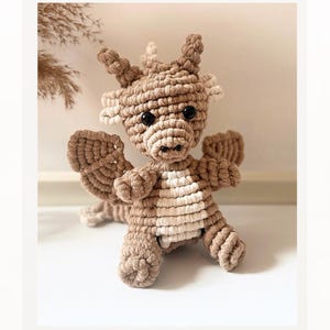May include: A handmade macrame dragon in shades of brown and cream. The dragon has large black eyes, small horns, and detailed wings. The body is a mix of brown and cream-colored macrame knots, creating a textured appearance. The dragon is sitting upright.