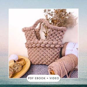 May include: A light brown macrame bag with a woven handle and a row of knotted balls around the top. The bag is sitting on a grey and white textured surface with a ball of light brown cord and a wooden spool of cord next to it.