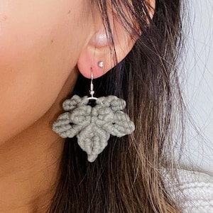 May include: Grey crocheted leaf-shaped earrings with silver hooks. The earrings are hanging from a person's ear.