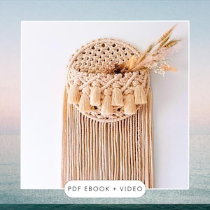 以下が含まれることがあります： 円形のデザインとタッセルが付いたベージュのマクラメ壁掛け。壁掛けの上部には小さなポケットがあり、ドライフラワーが入っています。画像の下部には「PDF EBOOK + VIDEO」というテキストがあります。