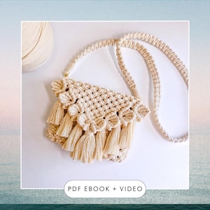 Op de afbeelding: Crèmekleurige macramé crossbodytas met een geweven ontwerp en lange kwasten. De tas heeft een lange riem en een decoratieve geschulpte rand. De tekst "PDF EBOOK + VIDEO" staat in de hoek. Een bol garen staat op de achtergrond.