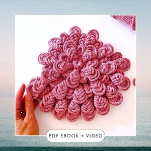 Puede incluir: Un gran tapiz de macramé circular de color rosa con un diseño texturizado único. El tapiz está hecho de cuerda gruesa y presenta una serie de nudos superpuestos que crean un efecto 3D. El texto "PDF EBOOK + VIDEO" está impreso debajo del tapiz.