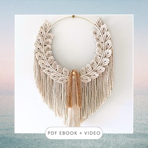 May include: A macrame wall hanging with a circular hoop and a fringe of tassels. The macrame is cream coloured and features a leaf pattern. The text "PDF EBOOK + VIDEO" is at the bottom of the image.
