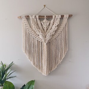 Peut inclure: Une grande suspension murale en macramé de couleur blanc cassé avec un motif géométrique. La suspension est réalisée avec un cordon de coton naturel et comporte une baguette en bois en haut.