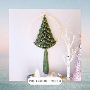 May include: A green yarn Christmas tree wall hanging with gold beads and a wooden hoop. The tree has a long green tassel hanging from the bottom. The hanging is on a white wall with a small white tree and a birch tree in the background. The text "PDF EBOOK + VIDEO" is at the bottom of the image.