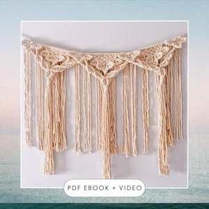 May include: A macrame wall hanging with a cream colored, geometric design. The hanging has a series of tassels along the bottom. The text "PDF EBOOK + VIDEO" is at the bottom of the image.