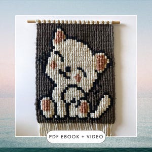 Może przedstawiać: Wiszący obraz z brązowej i białej przędzy z pikselowym wzorem kota. Kot jest biały z brązowymi oczami i różowymi policzkami. Obraz ma drewniany drążek u góry i frędzle u dołu. Pod obrazem znajduje się napis "PDF EBOOK + VIDEO".