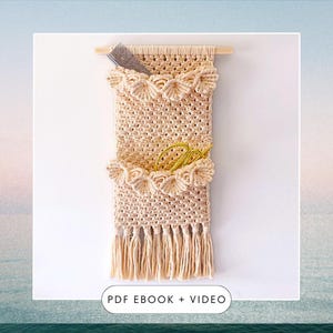 以下が含まれることがあります： ベージュの糸で編まれたマクラメの壁掛け。2つのポケットとフリンジの縁取りがあり、ポケットは波状の縁取りで飾られています。壁掛けは木製の棒にかけられています。