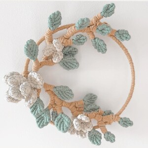 May include: A macrame wreath featuring a circular frame with beige rope. The wreath is adorned with light green leaves and off-white flowers, creating a natural and handcrafted aesthetic. The wreath is designed to be hung on a wall.