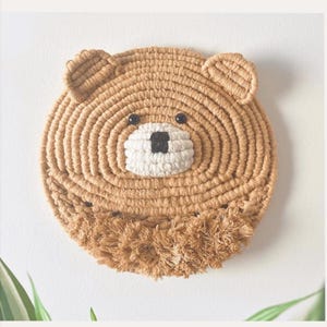 May include: A round, tan-colored wall hanging in the shape of a bear's face. The bear's face is made of woven rope, with black eyes, a white snout, and a black nose. The bear has small, rounded ears and a fringed area at the bottom.