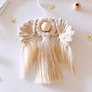 May include: A cream and beige macrame angel ornament. The angel has a wooden bead head, a halo, and a long tassel skirt. Gold star-shaped decorations are scattered around the angel on a white background. A wooden bead is also visible.