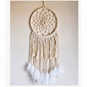 May include: A white macrame dream catcher with white feathers hanging from the bottom. The dream catcher is circular and has a intricate design.