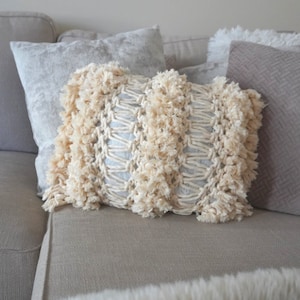 Op de afbeelding: Een crèmekleurig kussen met een geweven textuur en franjes. Het kussen ligt op een beige bank met andere kussens.