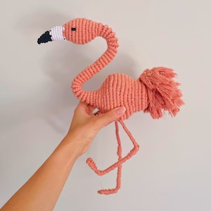 Puede incluir: Un flamenco rosa hecho de cuerda con un pico blanco y negro y una cola esponjosa. El flamenco está de pie sobre un fondo blanco.