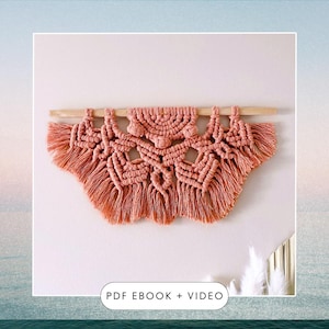 Op de afbeelding: Een perzikkleurige macramé wandhanger met een geometrisch ontwerp en franjes. De hanger is bevestigd aan een houten stok.