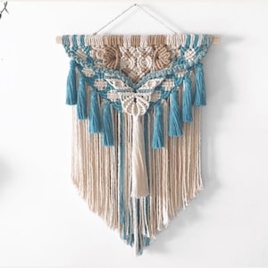 Puede incluir: Un tapiz de macramé en tonos beige y azul. Presenta una barra de madera, intrincados nudos y borlas colgantes. Esta obra de arte textil decorativa es ideal para añadir un toque bohemio a cualquier espacio, aportando un toque artesanal.
