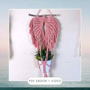 May include: A pink macrame plant hanger with a wooden dowel at the top. The hanger is holding a white pot with a green plant inside. The text "PDF EBOOK + VIDEO" is at the bottom of the image.