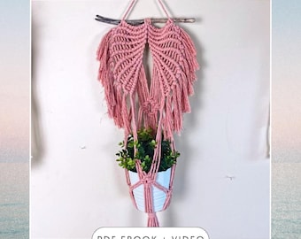 May include: A pink macrame plant hanger with a wooden dowel at the top. The hanger is holding a white pot with a green plant inside. The text "PDF EBOOK + VIDEO" is at the bottom of the image.