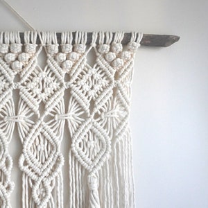 Pu&ograve; includere: Arazzo da parete in macram&egrave; bianco con un motivo geometrico. L'arazzo &egrave; realizzato con cordoncino di cotone ed &egrave; fissato a un bastone di legno.