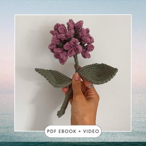 以下が含まれることがあります： ピンクの花の頭と緑の葉を持つ、かぎ針編みで作った花。花は手で持たれている。