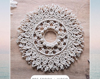 May include: A large, round macrame wall hanging with a circular opening in the center. The macrame is made with natural colored cord and has a fringe border. The text "PDF EBOOK + VIDEO" is printed below the macrame.