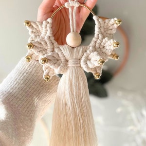 Puede incluir: Un adorno de ángel de macramé blanco con una cabeza de cuentas de madera y cascabeles dorados. El ángel tiene una larga borla que cuelga de la parte inferior.