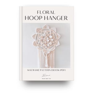 May include: A book cover featuring the title "FLORAL HOOP HANGER" in black. The image displays a cream macrame wall hanging with a floral pattern. The text "MACRAME PATTERN EBOOK (PDF)" is below the image, with a signature beneath.