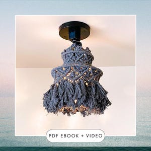 Op de afbeelding: Een grijze macramé plafondlamp met een zwarte metalen basis en franjes. De tekst "PDF EBOOK + VIDEO" wordt onder de lamp weergegeven.