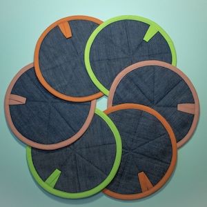 May include: Six denim-look round trivets with colorful trim in orange, green, and peach. Each trivet has a stitched radial pattern and a small tab. The trivets are arranged in a flower-like pattern.